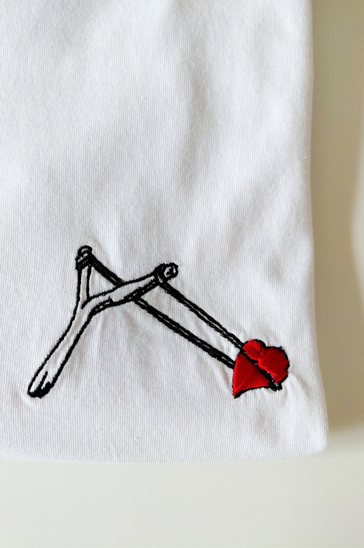 Close-up of embroidered slingshot heart design on white 100% cotton graphic t-shirt, quirky South African tee