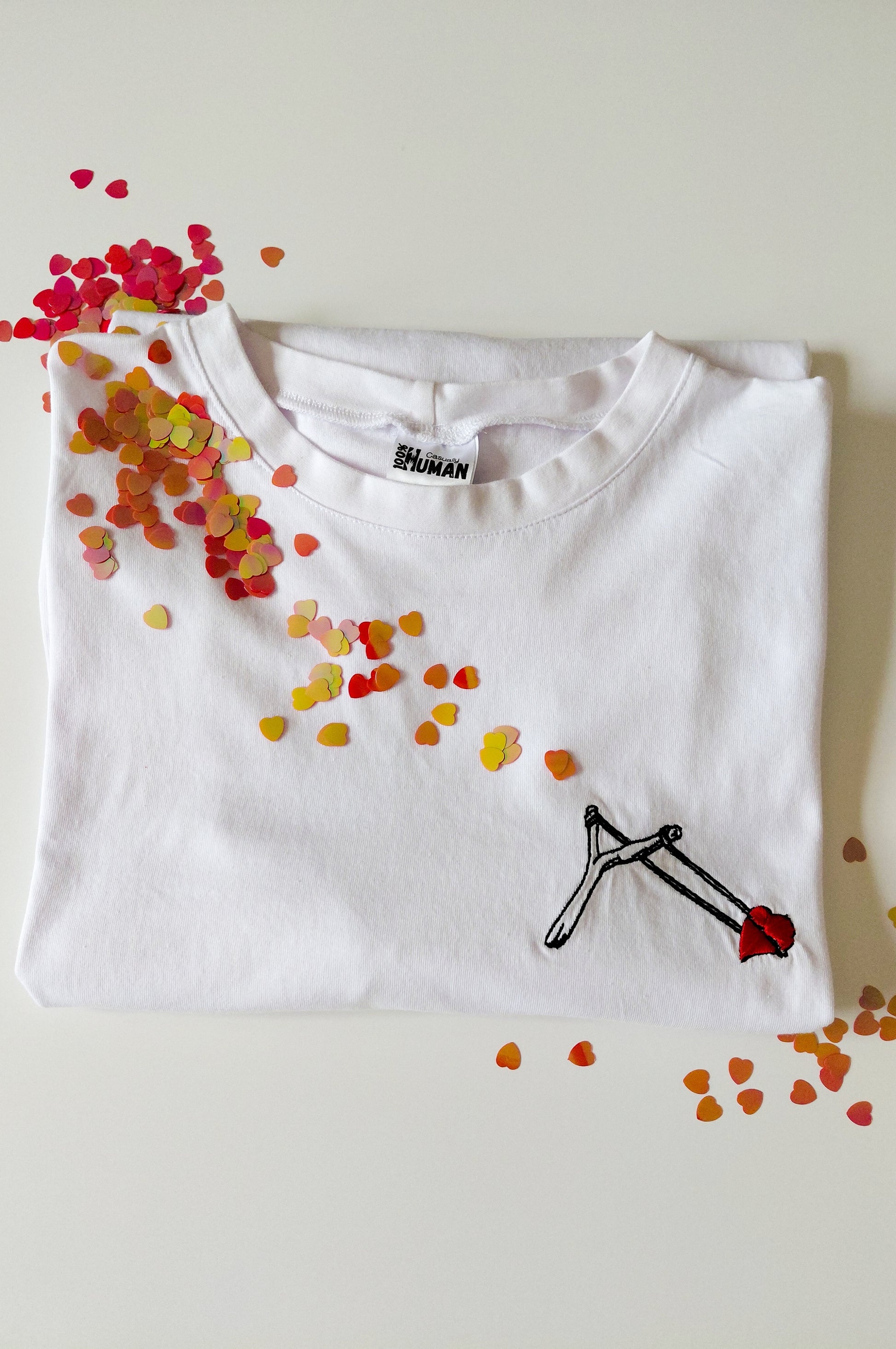 White graphic t-shirt with embroidered slingshot heart design, 100% cotton quirky tee flat lay South Africa