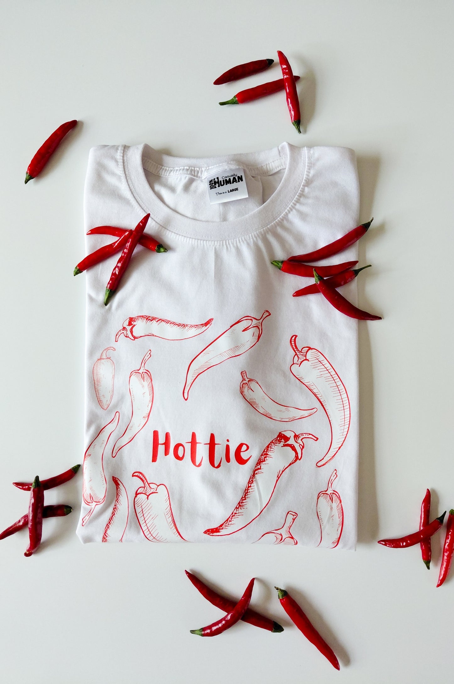 White graphic t-shirt with chilli print and Hottie text, 100% cotton funny tee flat lay with red chillies South Africa