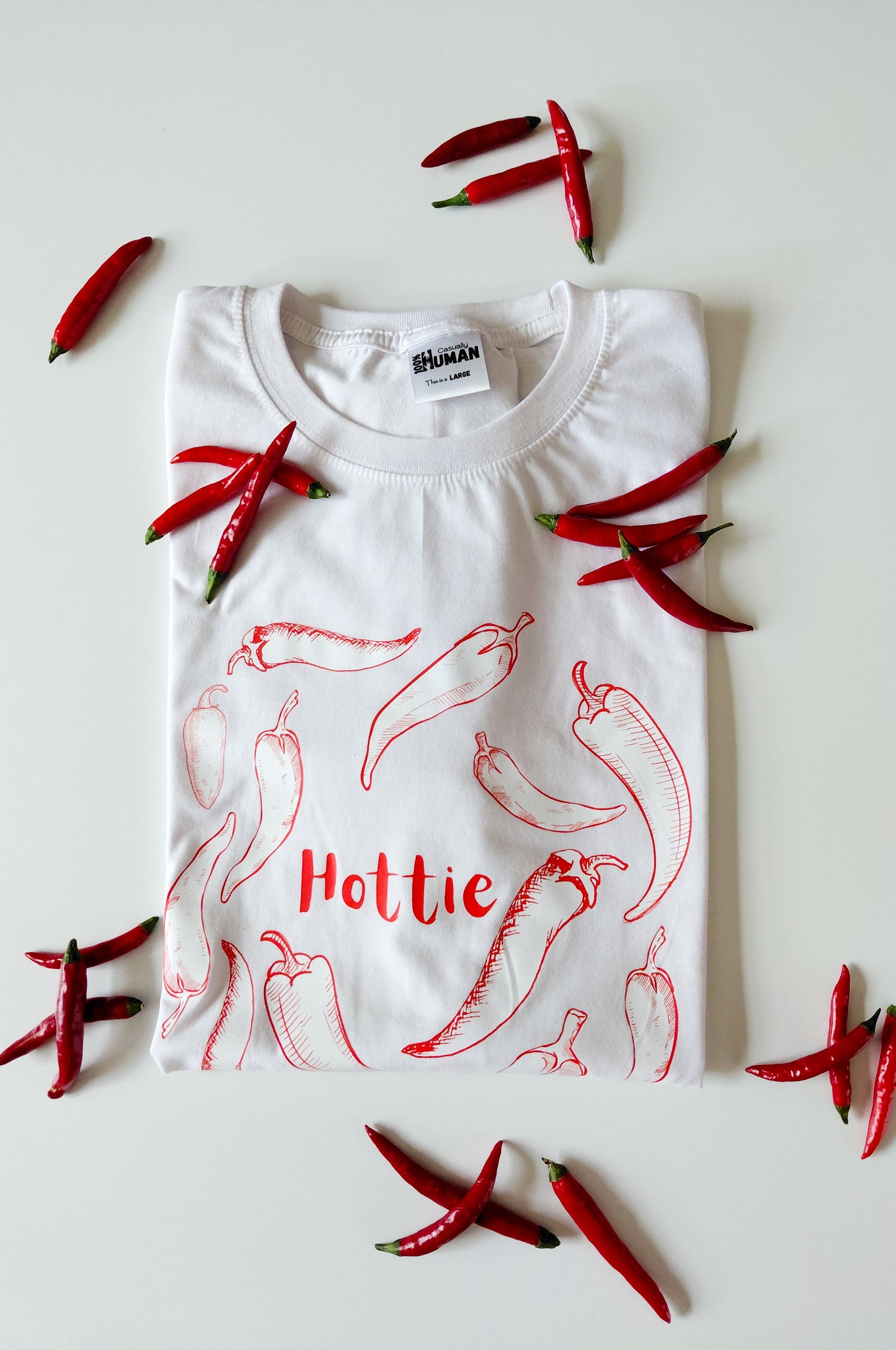 White graphic t-shirt with chilli print and Hottie text, 100% cotton funny tee flat lay with red chillies South Africa